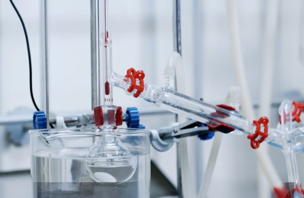 Close-up of an organic chemistry distillation setup showing flasks, a condenser and a thermometer connected with standard taper ground-glass joints secured by red Keck clips over a water bath.