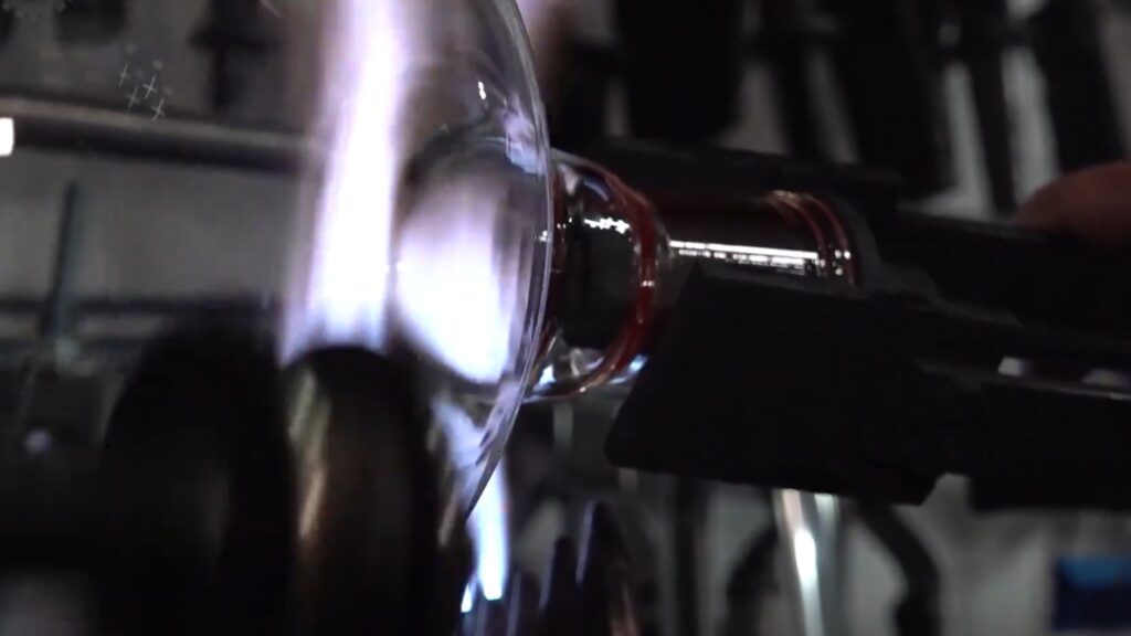 Close-up of a scientific glassblower heating a borosilicate flask over a blue torch, with the glowing standard taper ground joint held in a graphite clamp.